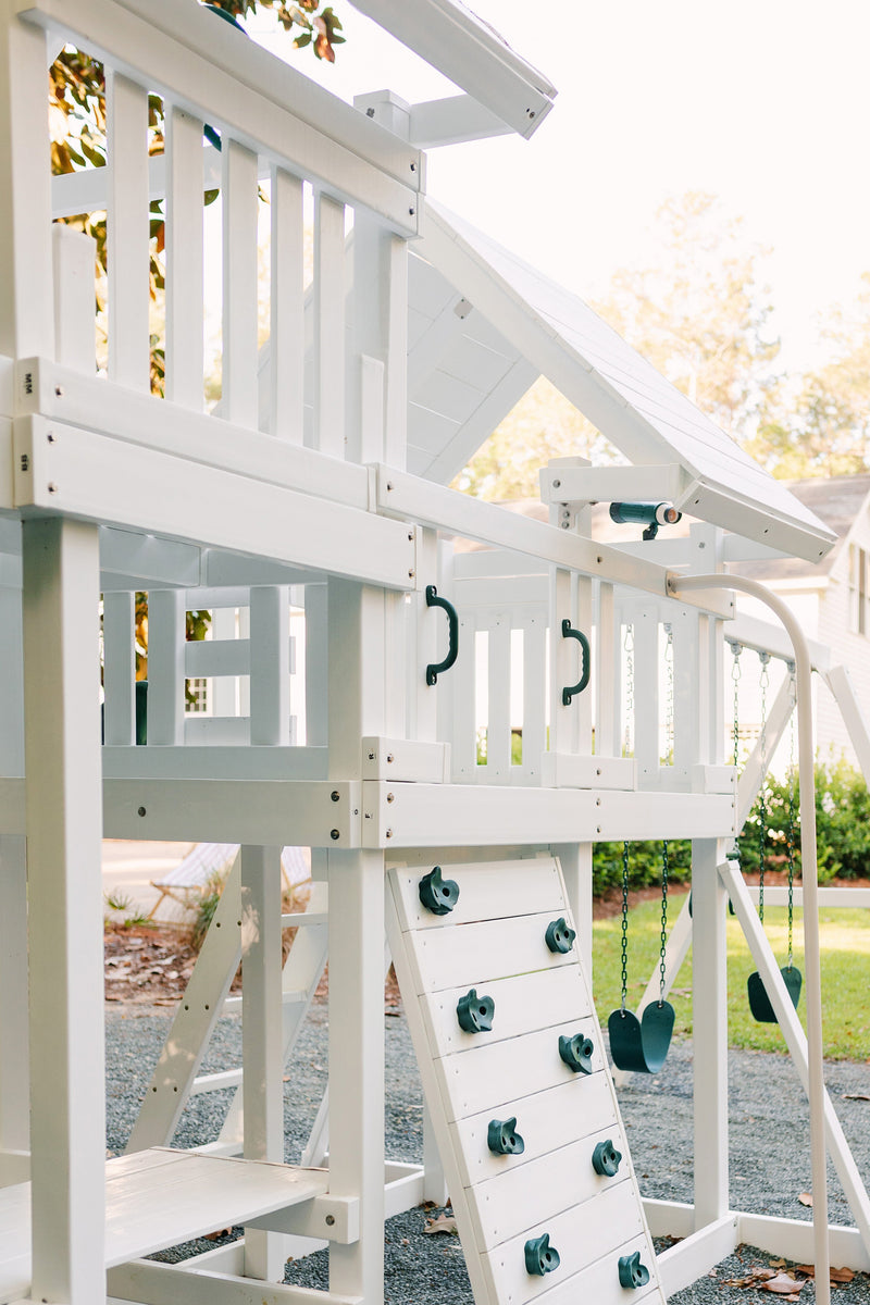 A large white wooden outdoor playset with a slide, climbing wall, rope swing, and elevated platform, photographed in a backyard setting on a gravel surface with grass and trees in the background.