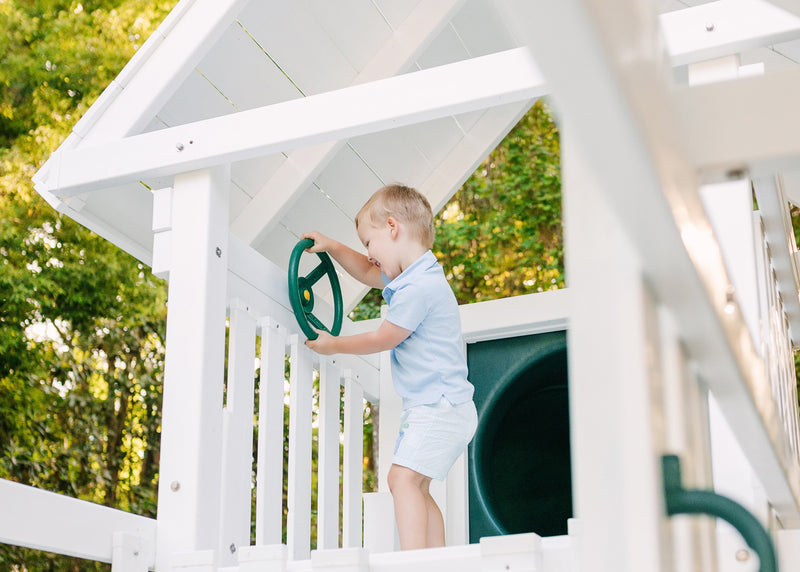 A young child in a light blue shirt and shorts stands on a white wooden playhouse, holding onto a green wheel handle. The scene is set outdoors with green trees in the background.