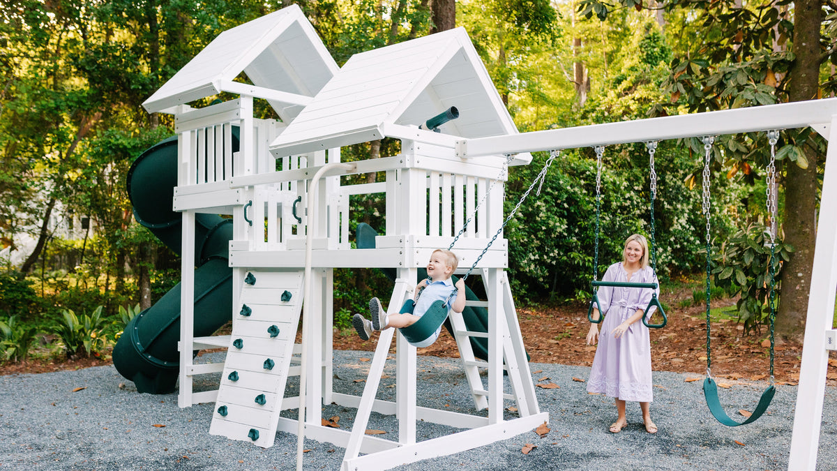 A large white wooden backyard playset with a spiral slide, climbing ladder, and swing set, set in a wooded backyard. A baby is sitting on the central swing, and a woman in a light pink dress stands nearby on the gravel ground.