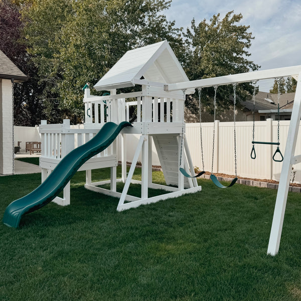 A white wooden backyard playset with a green slide, a small climbing platform, a swing set, and hanging rings, installed on a green lawn in a backyard with a white fence and trees.